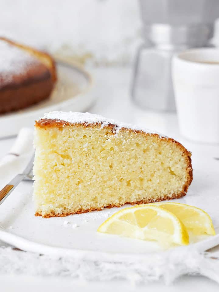 White plate with ricotta cake slice. A fork, lemon slices, white background with whole cake.