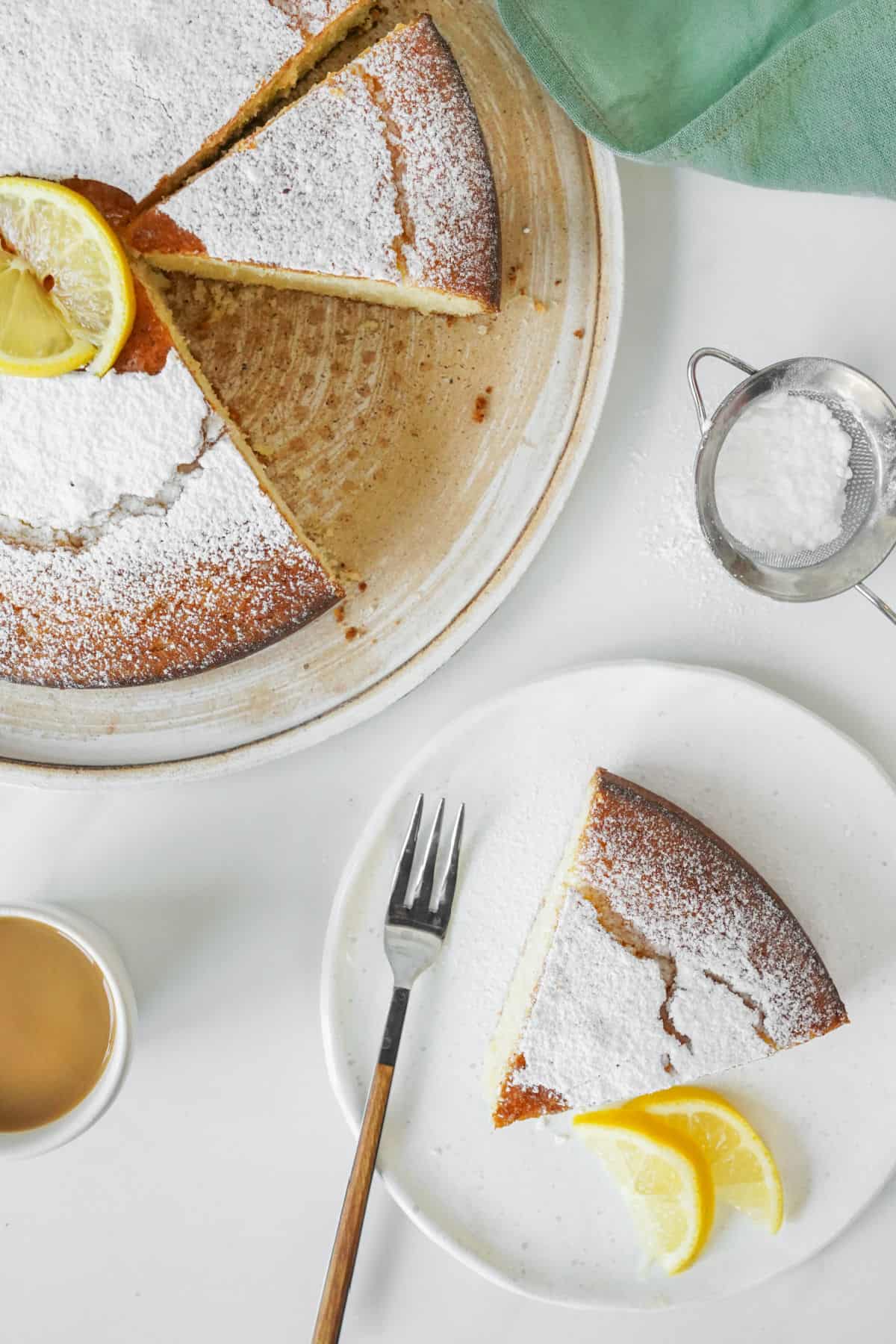 Top view of whole and slice of ricotta cake with powdered sugar. White surface, a fork, lemon slices.