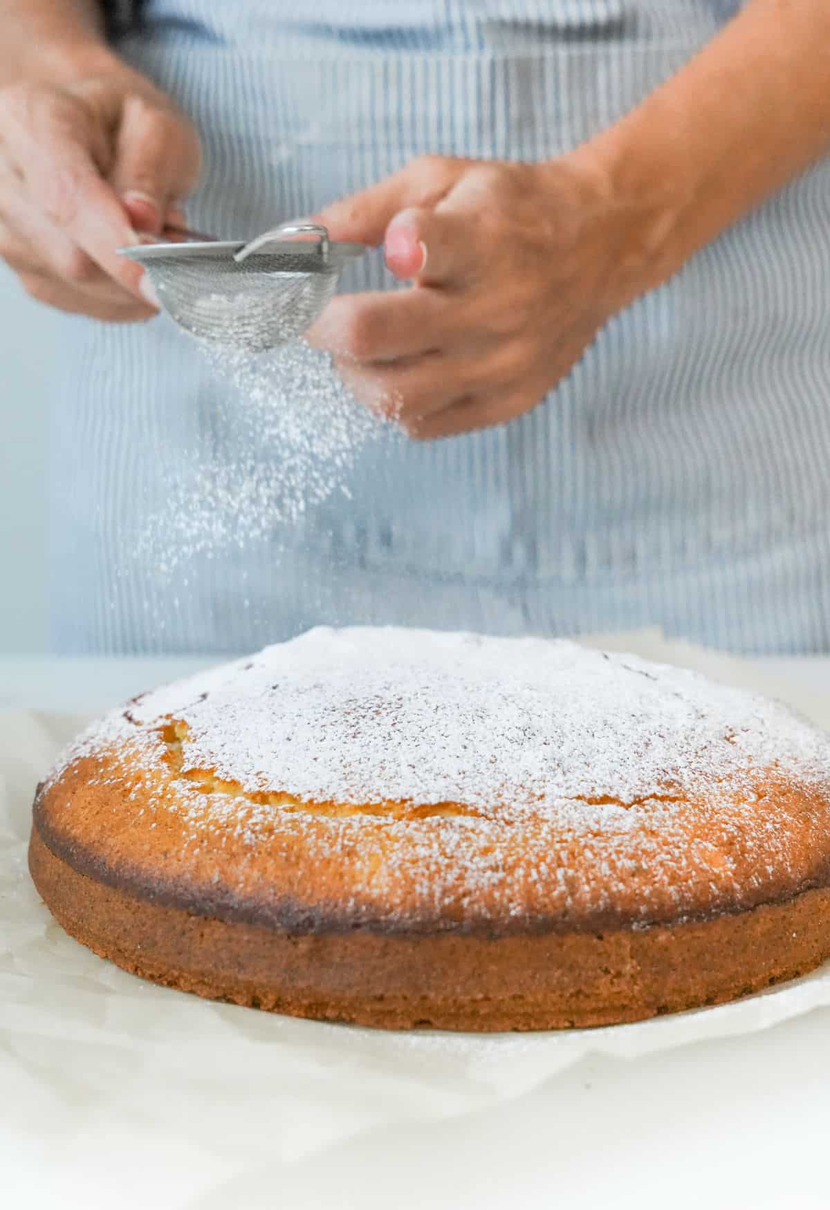 Sifting powdered sugar on plain ricotta cake. White surface. Striped apron background.