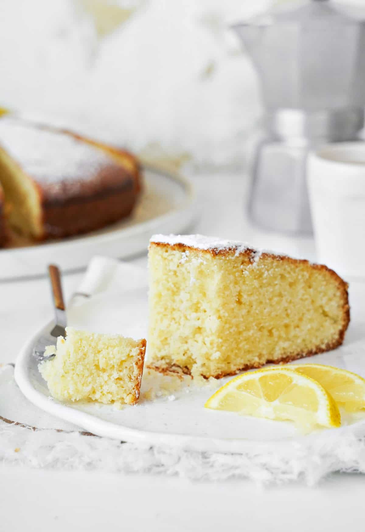 Slice of ricotta cake with forked bite on a white plate. White background. Lemon slices.