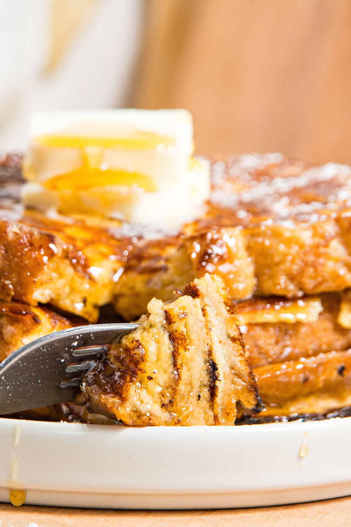 Close up of forked French toast bite with butter and syrup. White plate, wooden background.