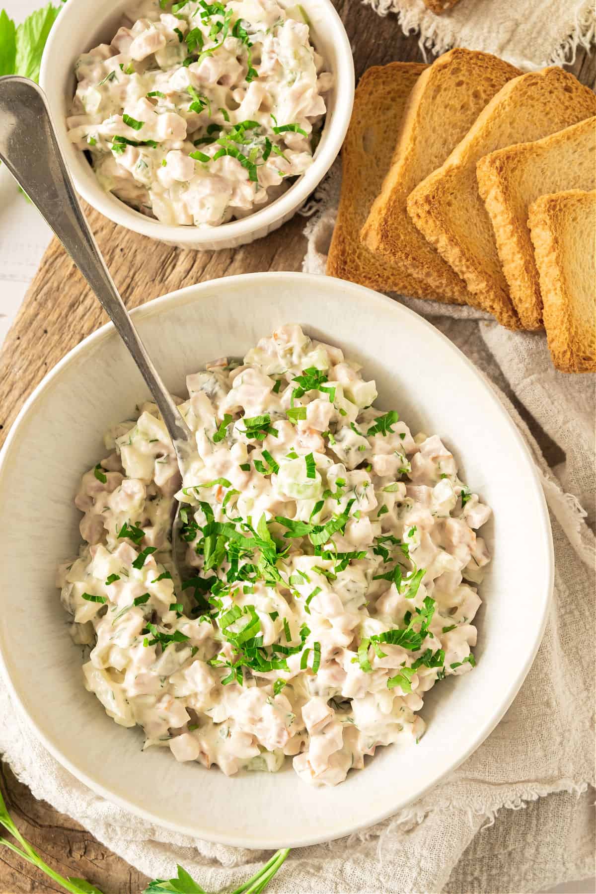 Top view of two white bowls with ham salad with chopped parsley. Beige cloth, toast slices.