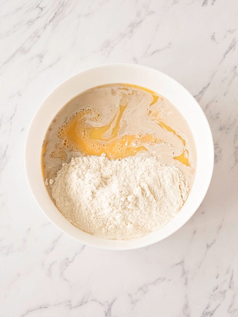 Flour and wet ingredients for bread in a white bowl. Top view. White marbled surface.