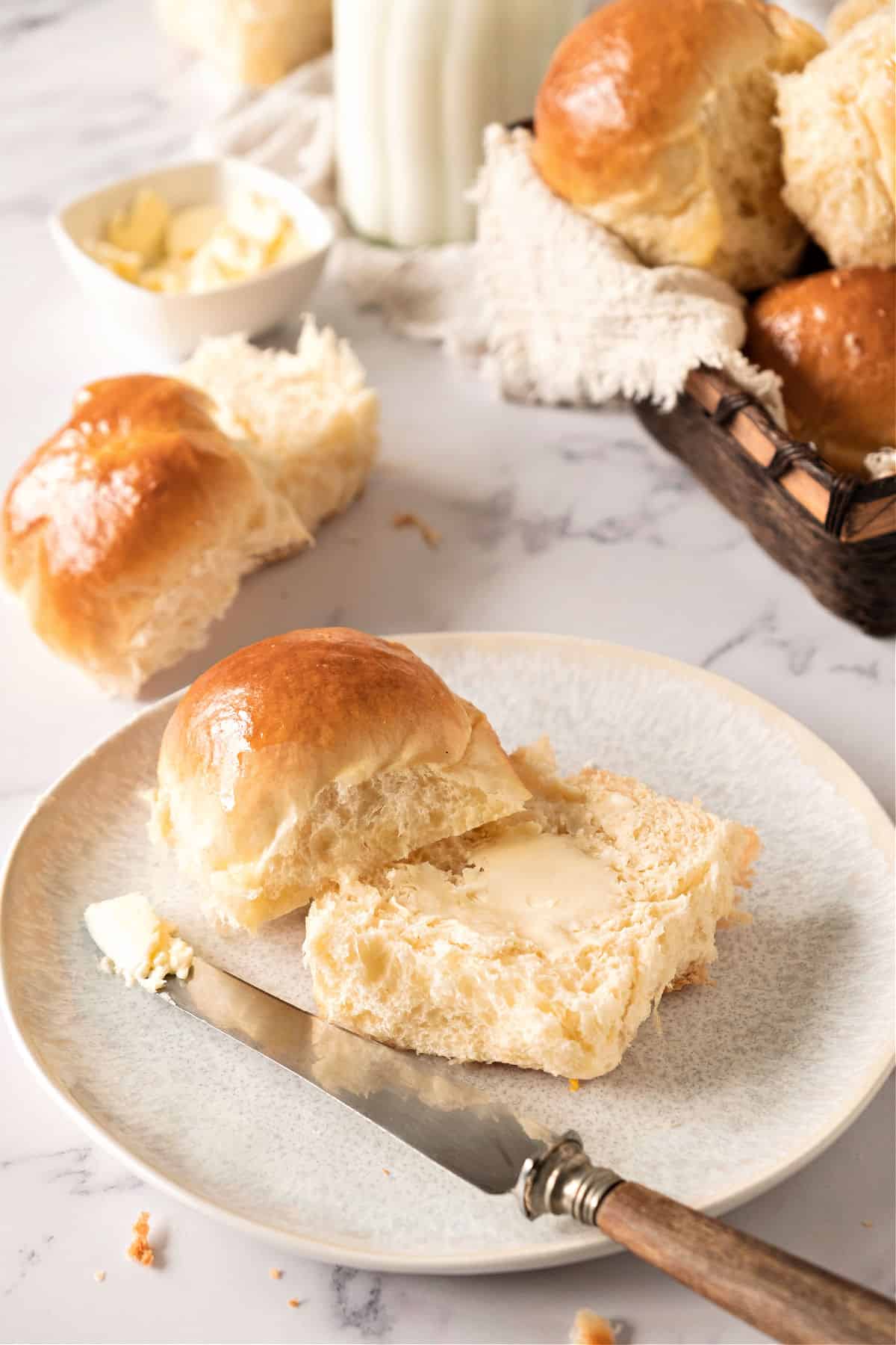 Halved soft roll with butter. White plate, a knife. White marble surface with more rolls.