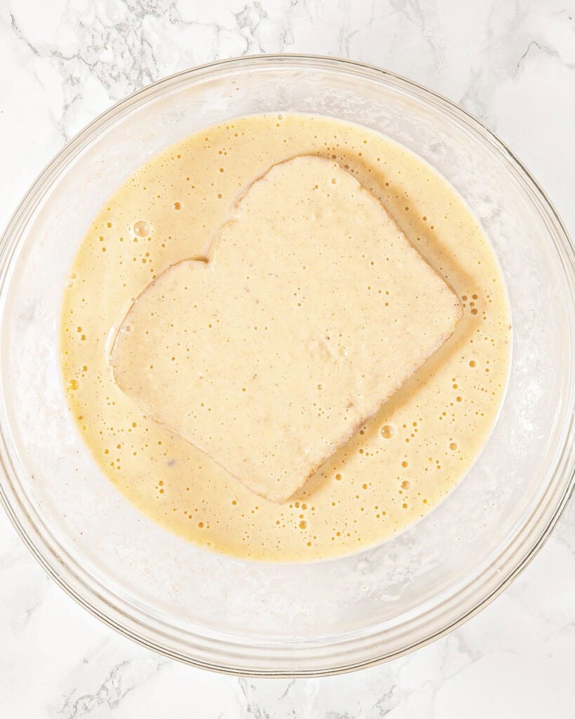 Soaking bread slice in eggnog mixture in a glass bowl. Top view. White marble.