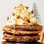 Red, brown and green text overlay on stack of gingerbread pancakes with whipped cream.