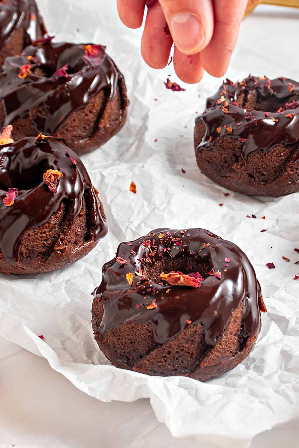 Sprinkling rose petals on chocolate glazed mini bundt cakes. White paper surface.