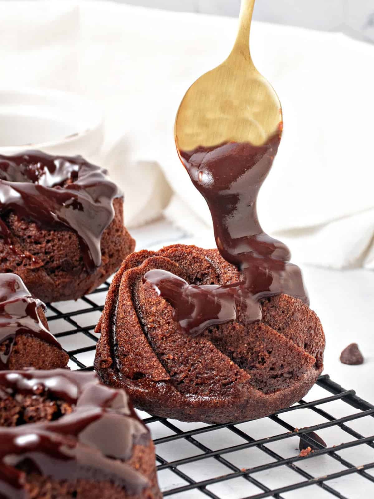 Glazing chocolate mini bundt cakes with ganache. Golden spoon, wire rack. White background. 