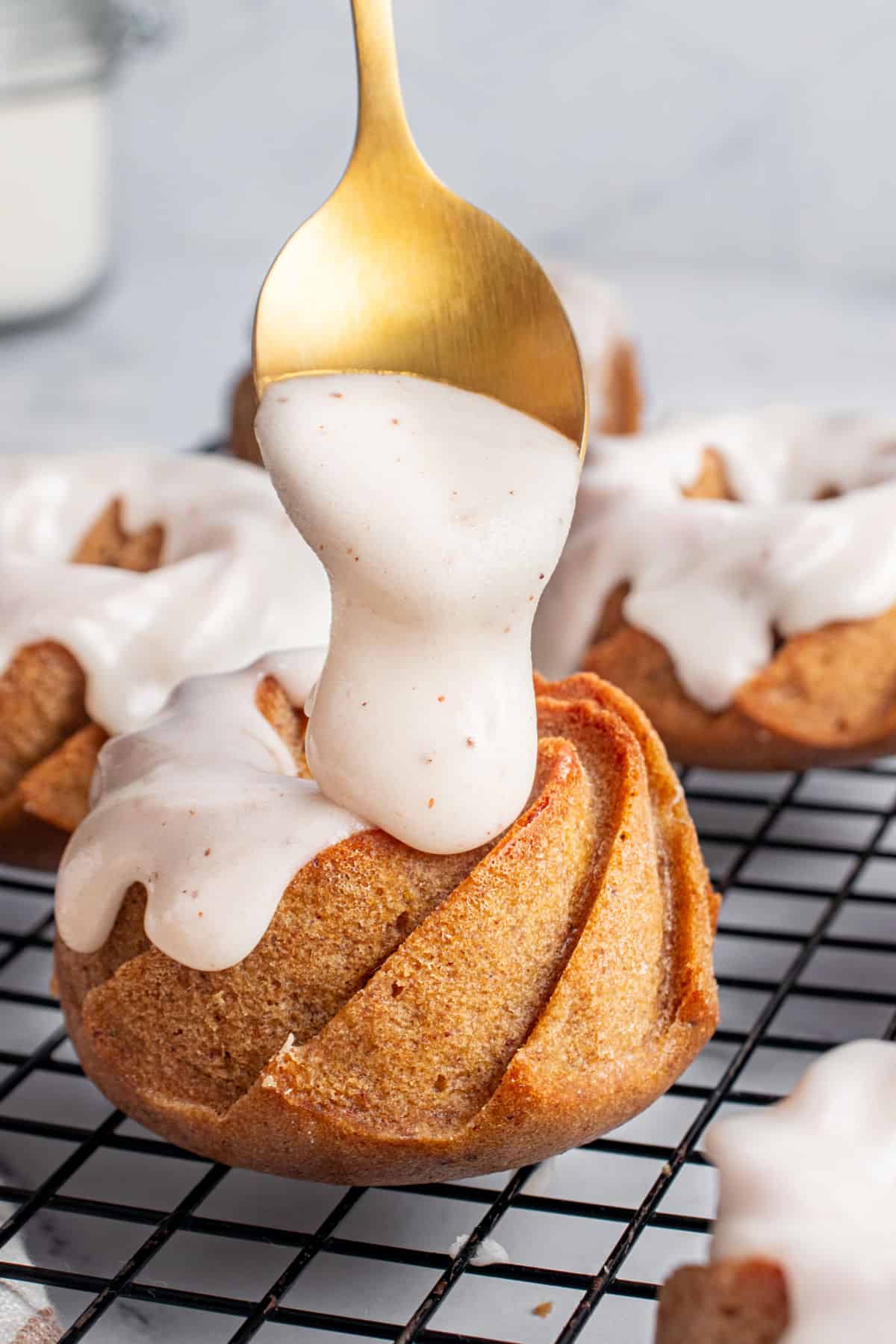 Glazing mini bundt pumpkin cake on a black wire rack. Golden spoon, gray background.