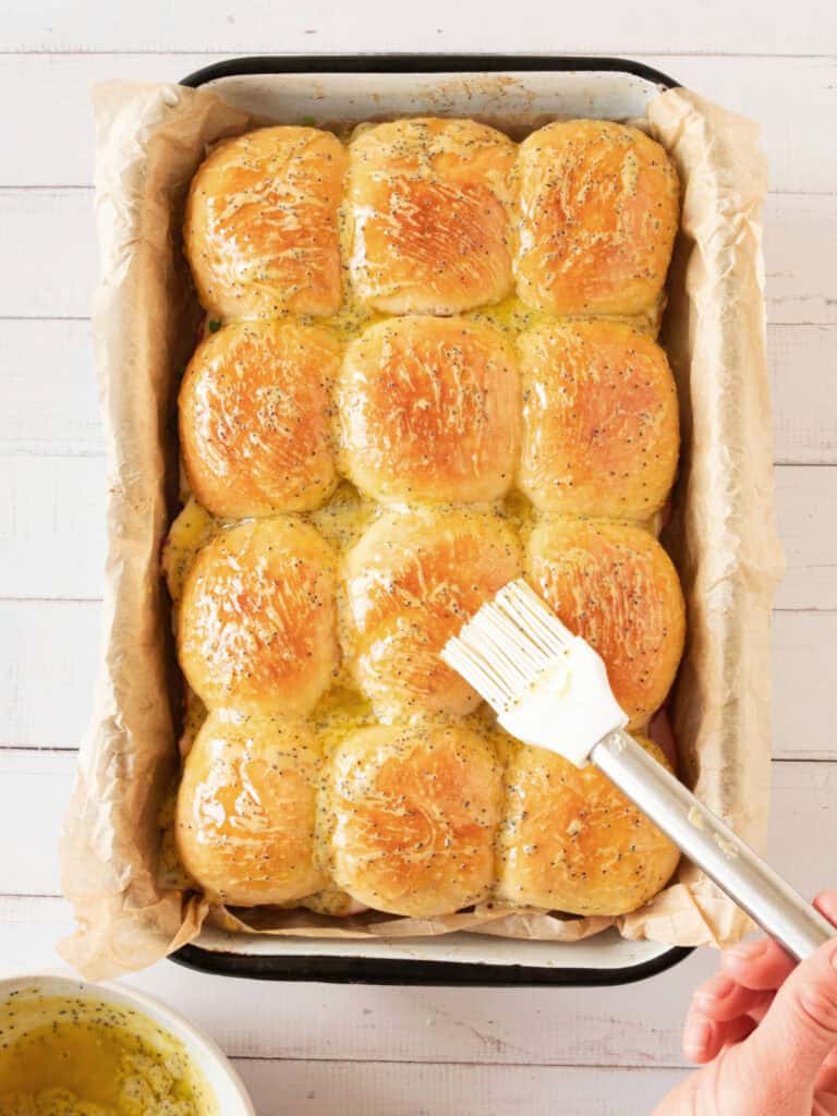 Brushing flavored melted butter on rows of small sandwiches in a baking dish. White background. Top view.