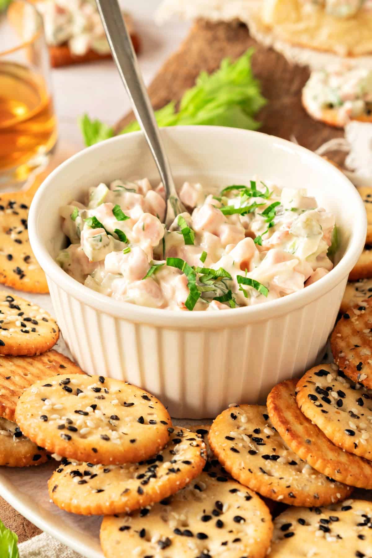 White bowl with ham spread. Silver spoon. Sesame crackers around.