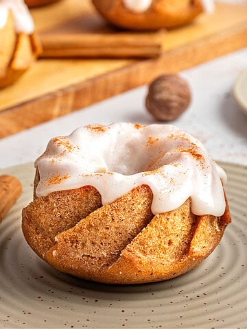 Dry green plate with single glazed mini pumpkin bundt cake. Wooden board, cinnamon sticks.