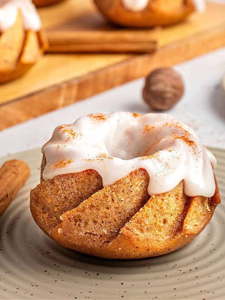 Dry green plate with single glazed mini pumpkin bundt cake. Wooden board, cinnamon sticks.