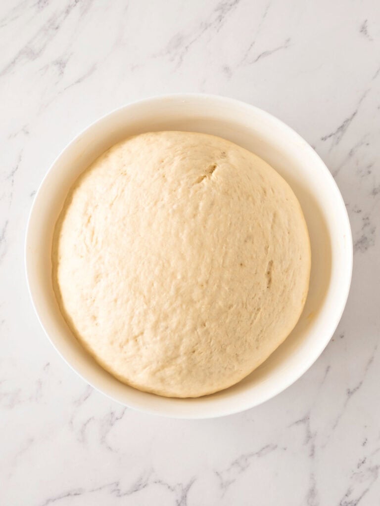 Doubled bread dough in a white bowl. Top view. White marbled surface.