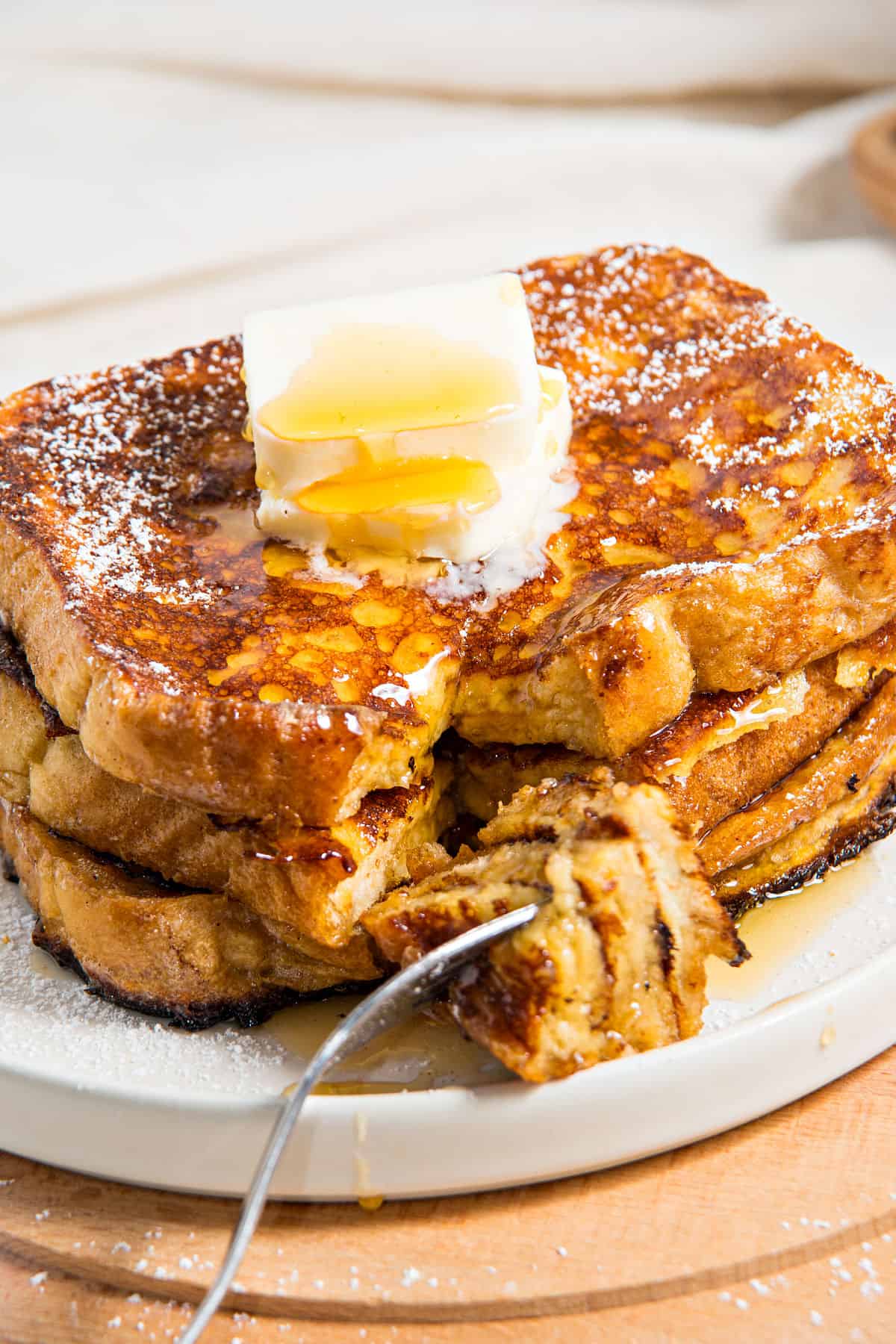 Fork with bite of French toast with whole serving on a white plate. Butter an syrup drizzle.