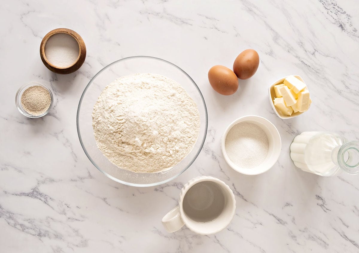 Ingredients for white dinner rolls in bowls on a white marbled surface.