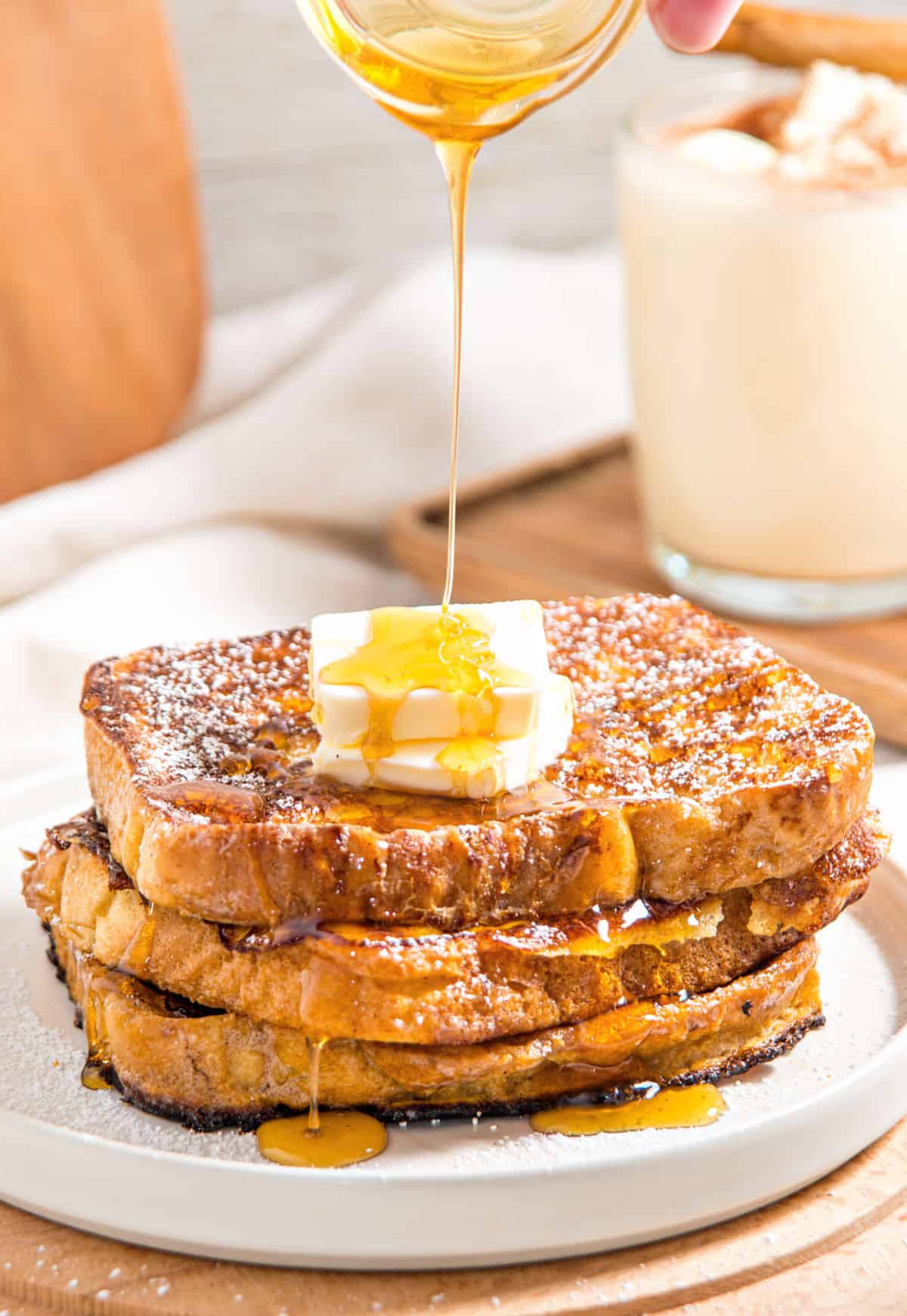 Pouring syrup onto French toast stack with butter. White plate, wooden surface, gray background.