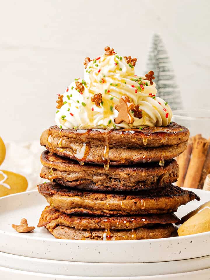 Stack of gingerbread pancakes with whipped cream and syrup on white plate. Christmas decorations, white background.