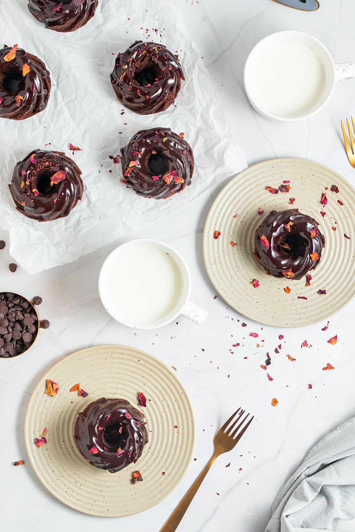 Top view of white surface with two plates and white paper with glazed chocolate mini bundts. Rose petals, forks, milk glasses.