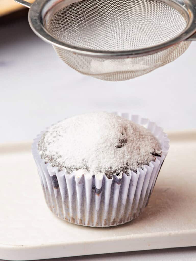 Sprinkling powdered sugar on a cupcake in a white paper liner. White background.