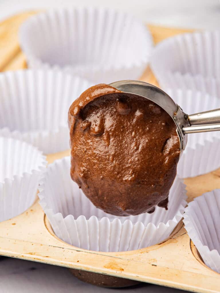 Scooping chocolate cake batter into white paper liners in light colored muffin tin.