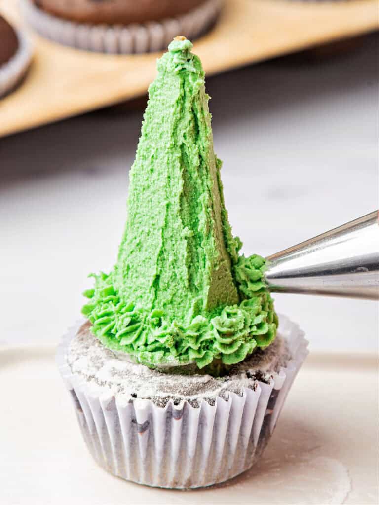 Piping green buttercream on frosted waffle cone atop a cupcake in a paper liner. White surface.