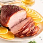 Sliced glazed ham on a white platter with orange slices and thyme.