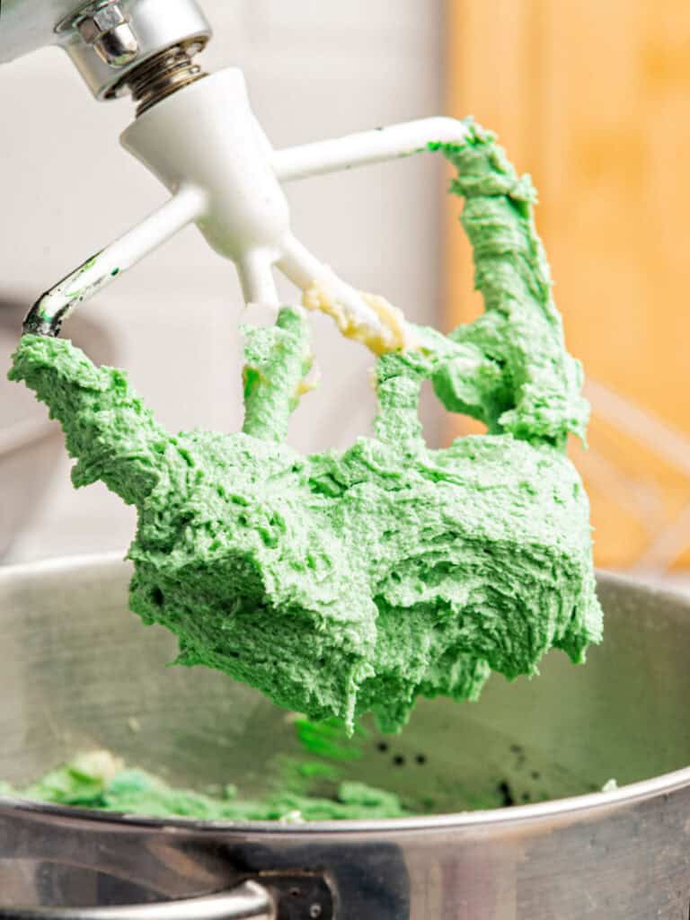 Stand mixer beater with green buttercream lifted from the metal bowl.