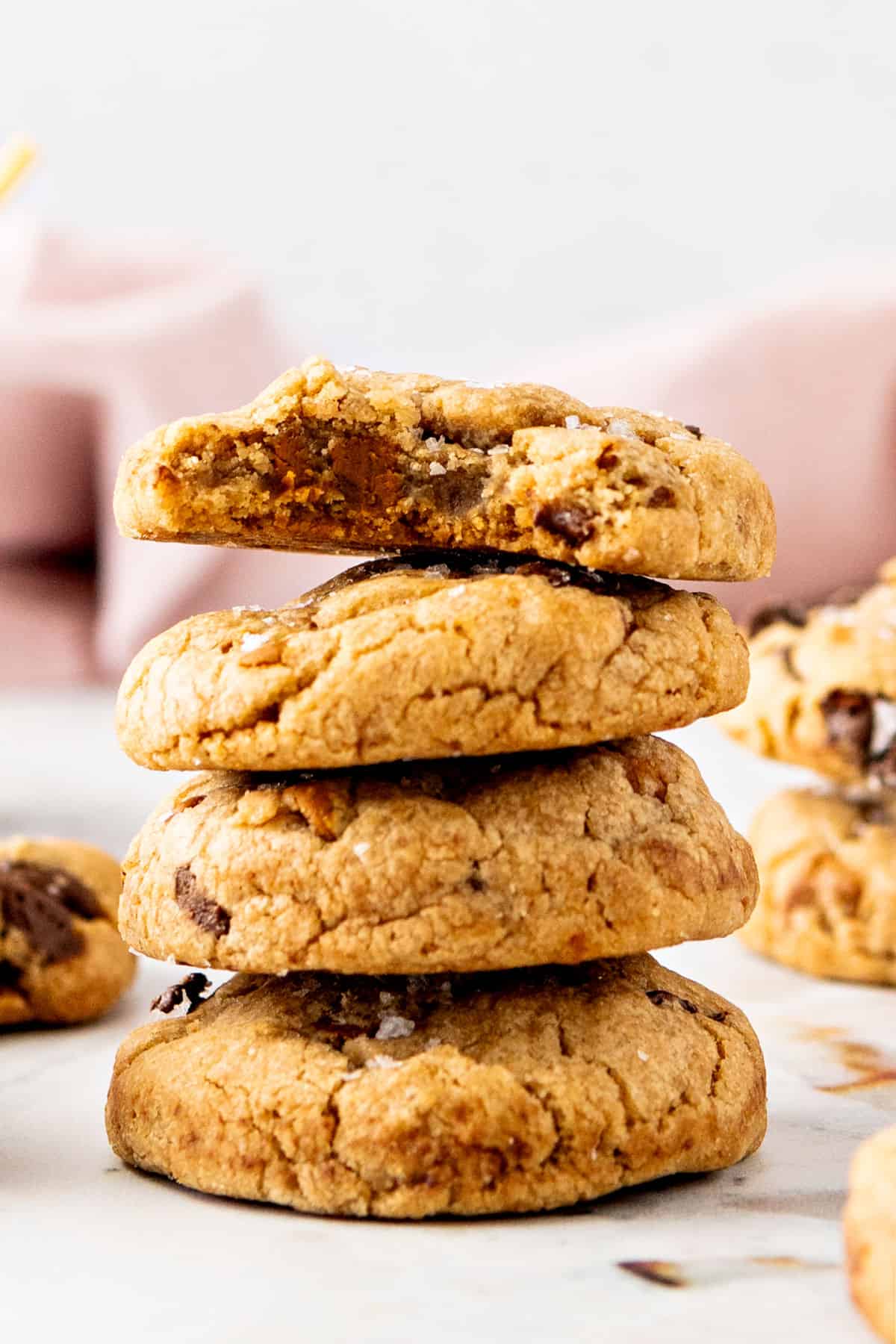 Four chocolate chip cookies in a stack, top one is bitten. White pink background.