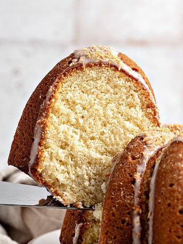 Lifting a slice of glazed coconut bundt cake from whole cake. Whitish tile background.