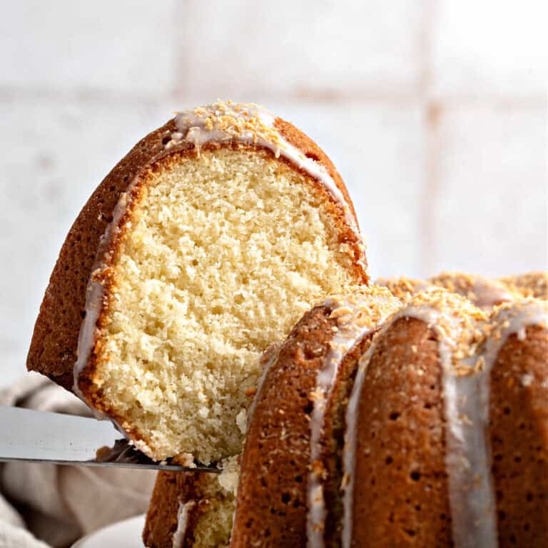 Lifting a slice of glazed coconut bundt cake from whole cake. Whitish tile background.