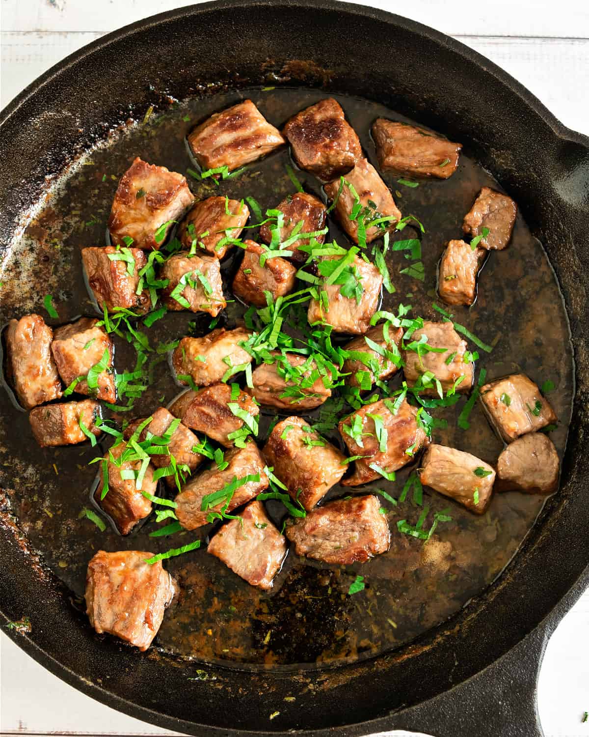 Parsley-topped steak bites in a cast-iron skillet. Top view.