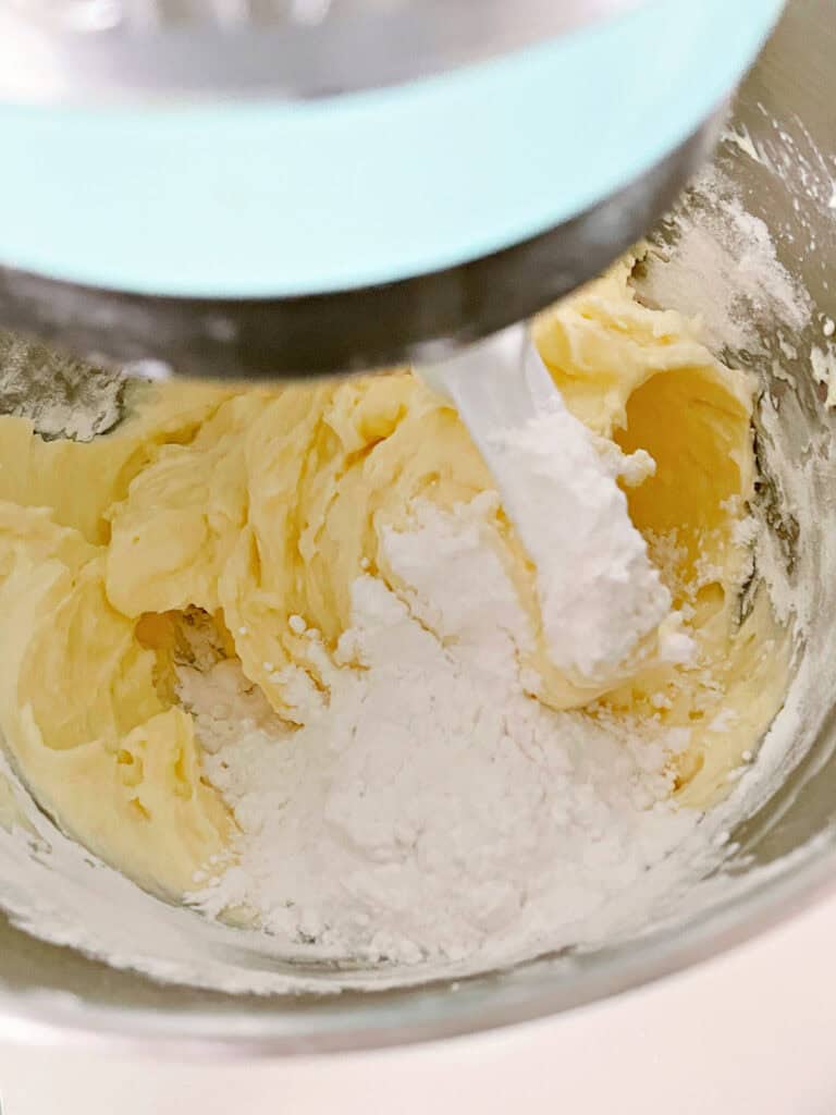 Powdered sugar added to butter mixture for frosting in a metal bowl. Stand mixer with paddle attachment.