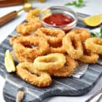 Gray marble board with pile of fried calamari. Bowl of ketchup, lemon wedges.