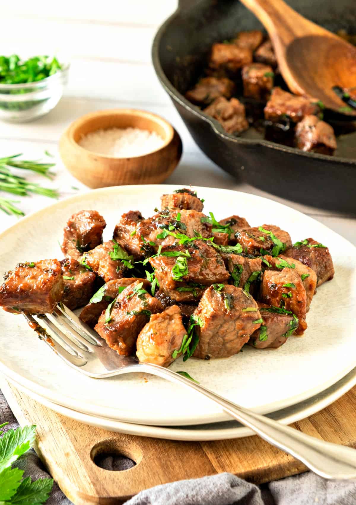 White plate with parsley steak bites, a silver fork. Cast iron pan in the white background.