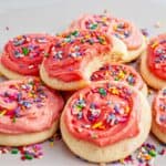 Pile of pink frosted sugar cookies with sprinkles. Top one is bitten. Gray background.