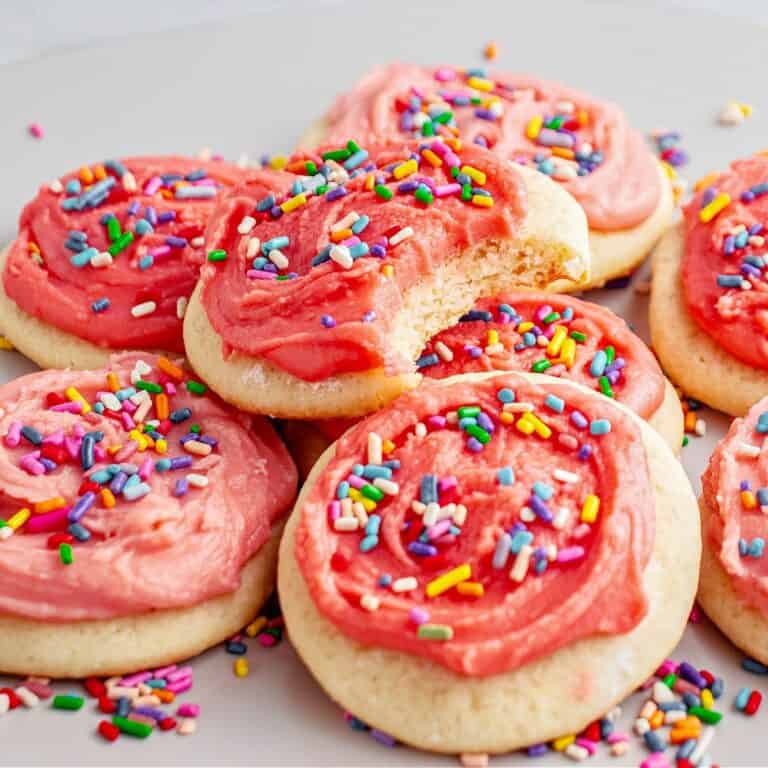 Pile of pink frosted sugar cookies with sprinkles. Top one is bitten. Gray background.