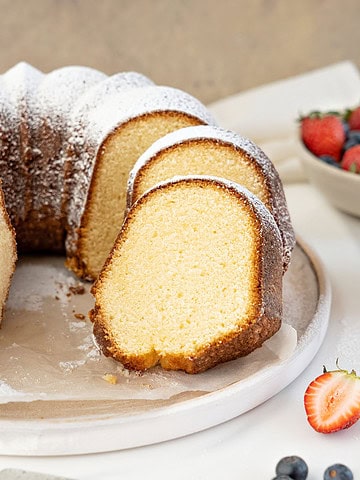 Whole and sliced plain bundt cake with powdered sugar on a white plate. Brown and white background.