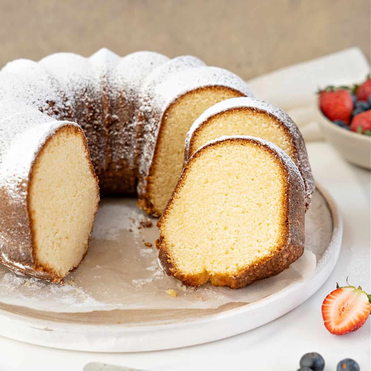 Whole and sliced plain bundt cake with powdered sugar on a white plate. Brown and white background.