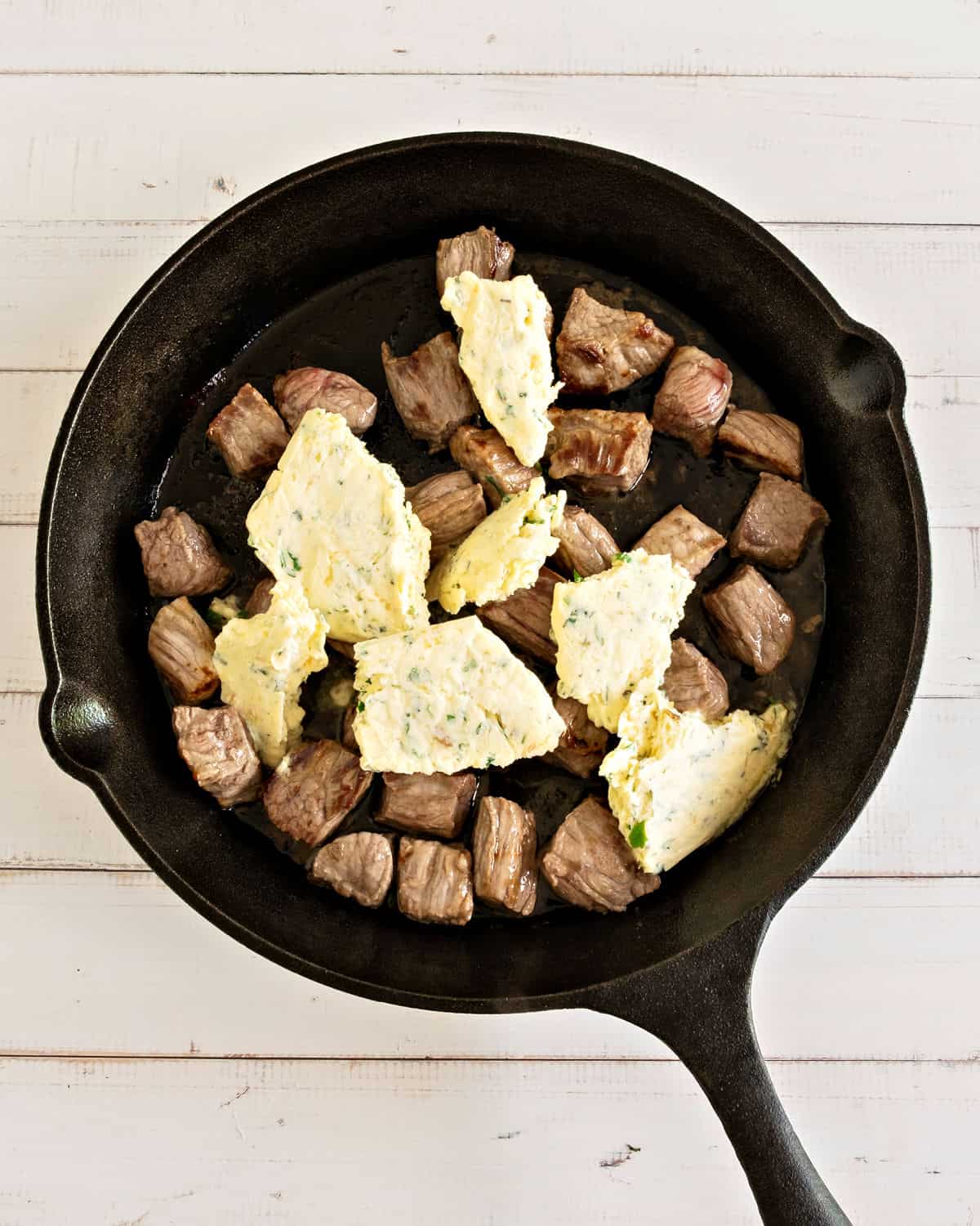 Cast-iron skillet with garlic butter pieces atop cooked steak bites. White background. 