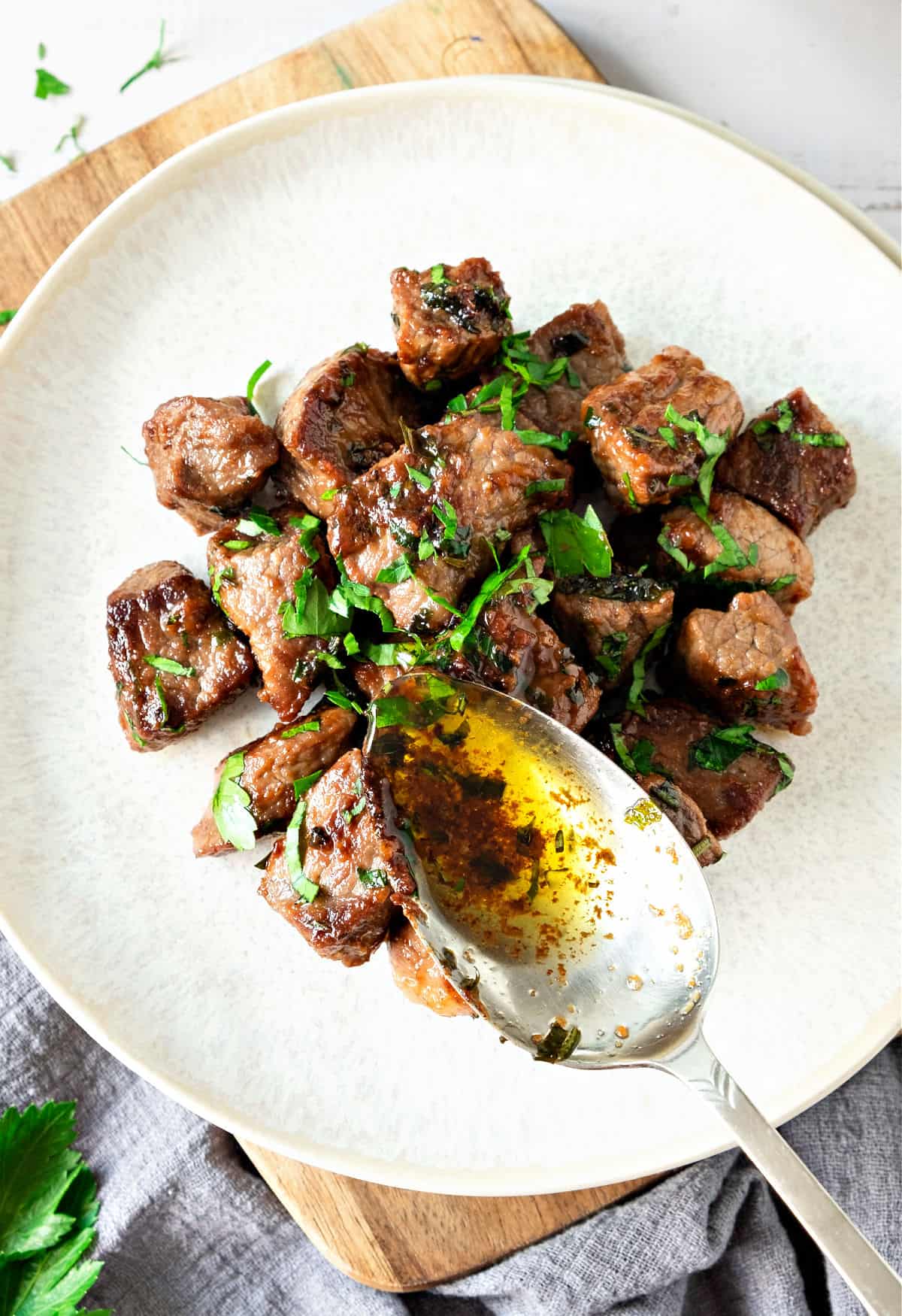 Pouring garlic butter sauce onto steak bites with parley on a white plate.