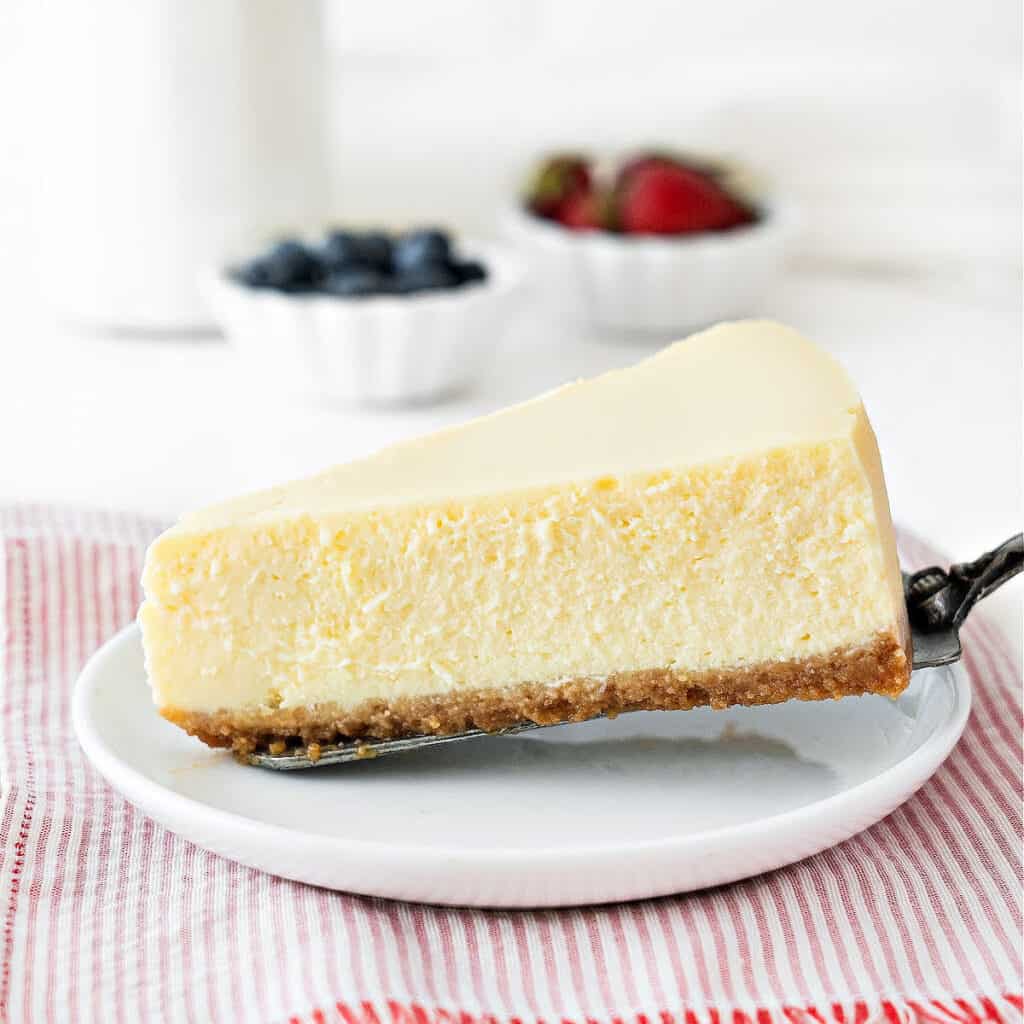 Close up slice of plain cheesecake on a white plate. Red striped cloth, bowls with berries, white background.
