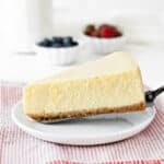 Close up slice of plain cheesecake on a white plate. Red striped cloth, bowls with berries, white background.