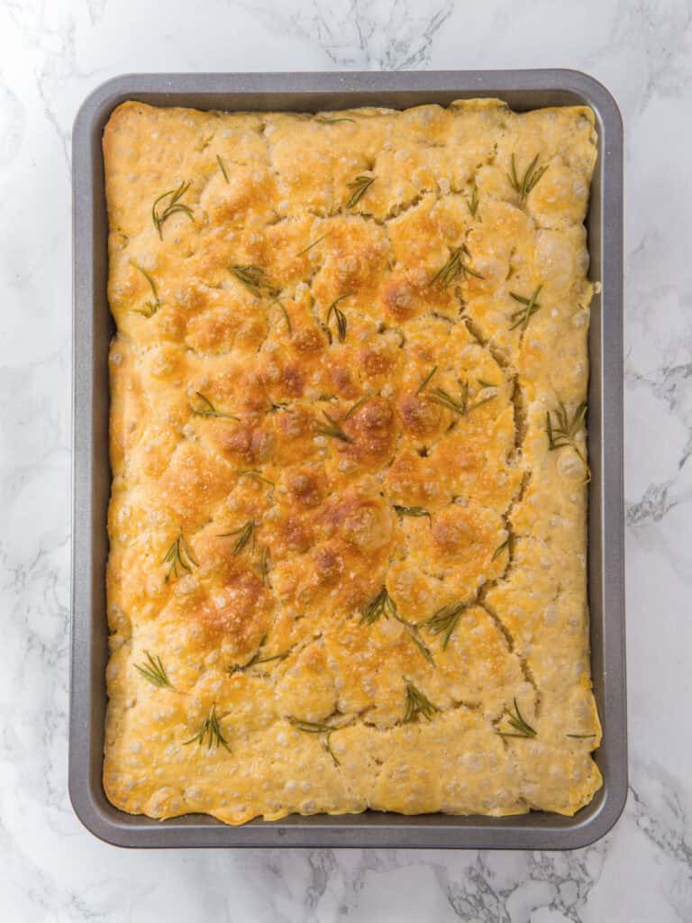 Top view of baked rosemary focaccia in a metal rectangular pan. White marbled surface.