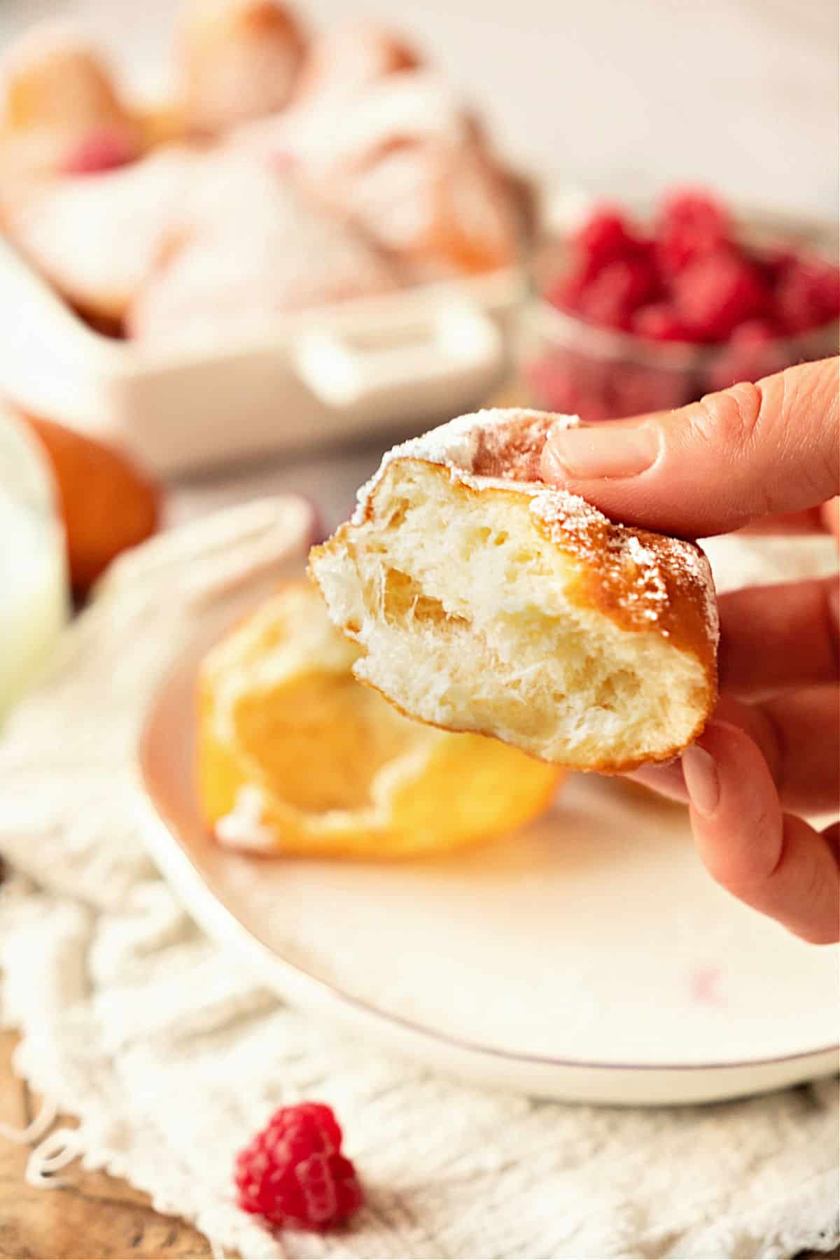 Bitten donut being held above white plates with more pastries, white cloth and raspberries.