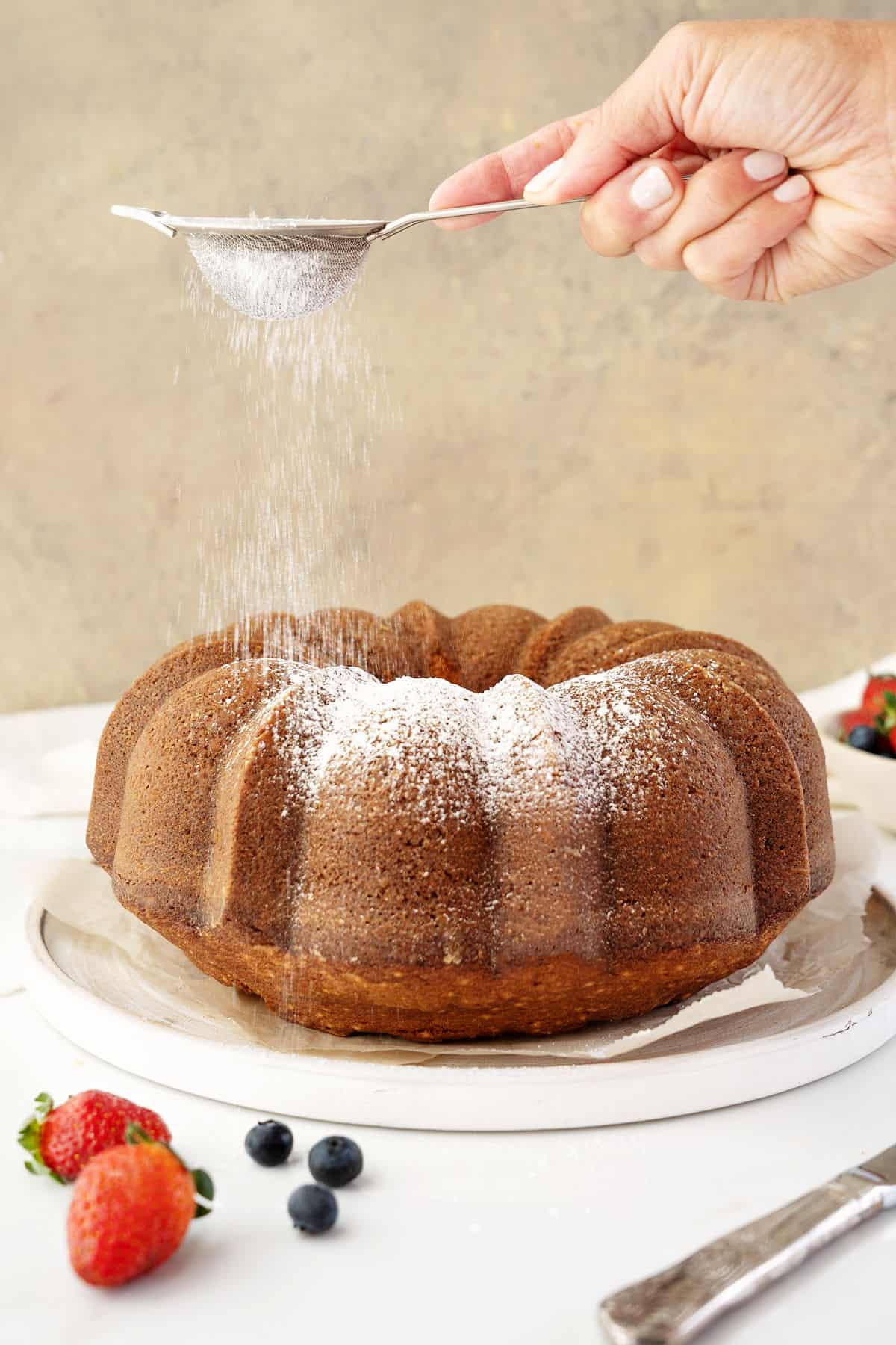 Dusting powdered sugar on a vanilla bundt cake on a white plate. Beige background, white surface, berries.