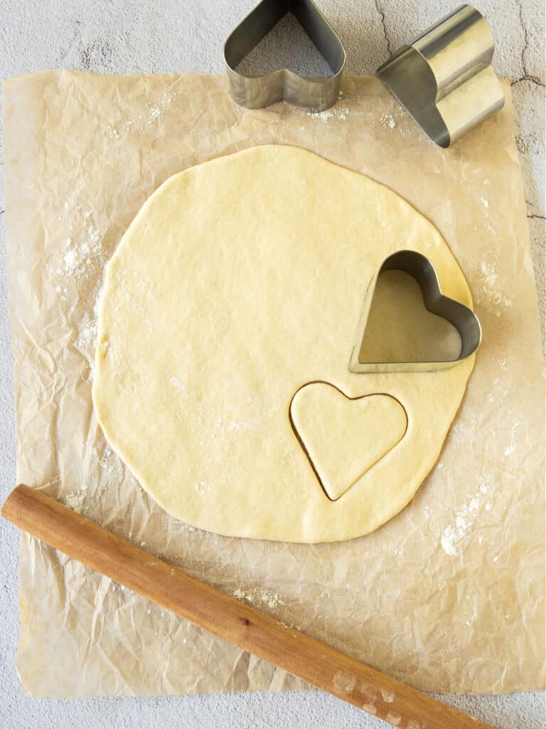 Rolled dough on parchment paper, rolling pin, heart shaped metal cutters. Top view.