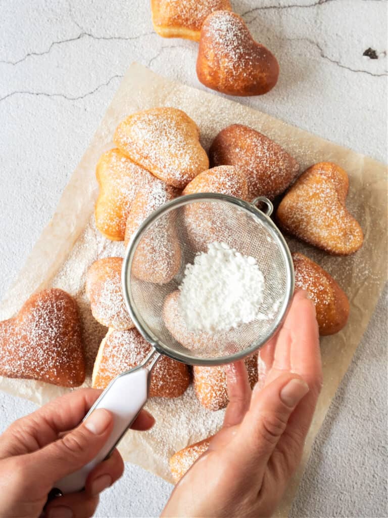 Sifting powdered sugar over pile of heart-shaped donuts on parchment paper. Gray background. Top view.