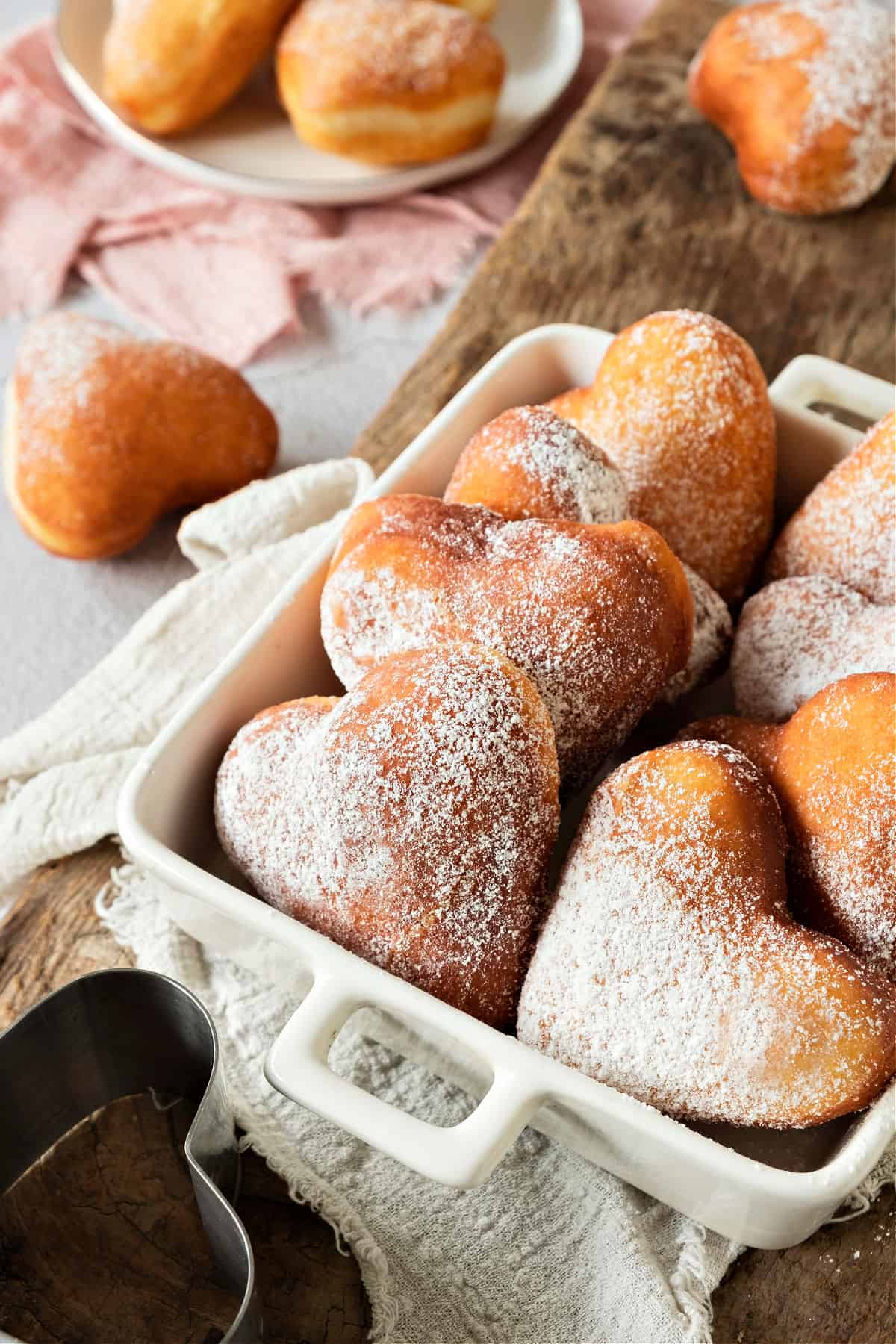 Heart-shaped donuts in a white baking dish. More on a plate in the background. Pink towel.