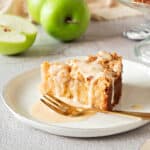 Slice of apple cake with custard sauce on a white plate. Gold fork, green apples in the light gray background.